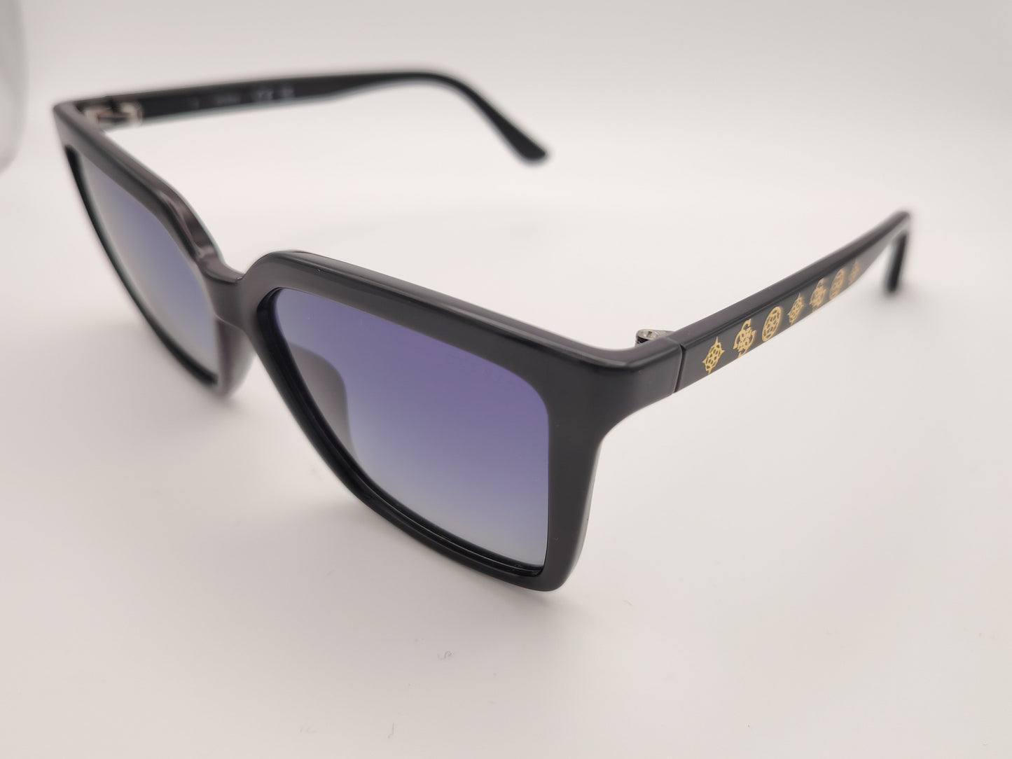 gafas de sol negras con montura en forma cuadrada, cristales degradados en tonos azulados y detalles dorados con símbolos decorativos en la varilla